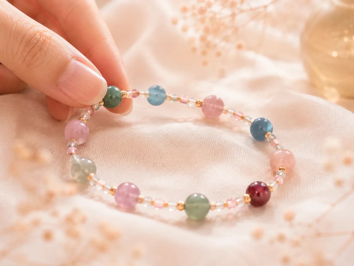 A hand lifting a pastel-bead bracelet from a soft pink fabric