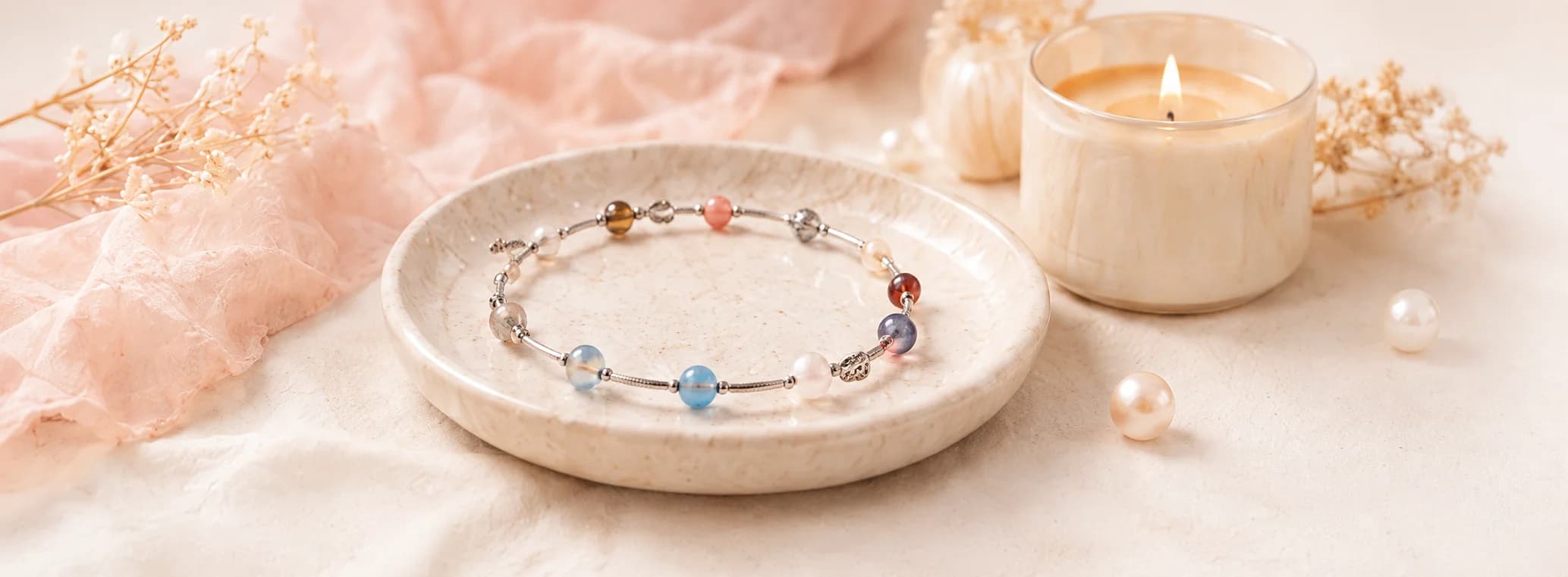 A multicoloured gemstone Lumoraah bracelet resting in a ceramic dish beside a lit candle, dried flowers, and loose pearls on soft pink linen